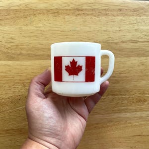Puede incluir: Taza de cerámica blanca con la bandera canadiense, con una hoja de arce roja sobre fondo blanco, enmarcada por barras verticales rojas. La taza tiene un asa curva y un aspecto ligeramente envejecido.