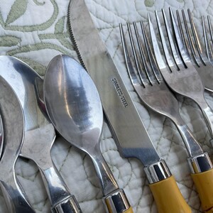 May include: Close-up of stainless steel silverware, including spoons, forks, and knives. The knives have serrated blades and yellow handles. The text "STAINLESS STEEL" is visible on the knife blade. The silverware is arranged on a patterned, off-white surface.