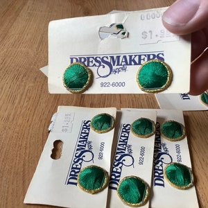 May include: Five green and gold embroidered buttons on white cards with the text "Dressmakers Supply" and the phone number "922-6000".