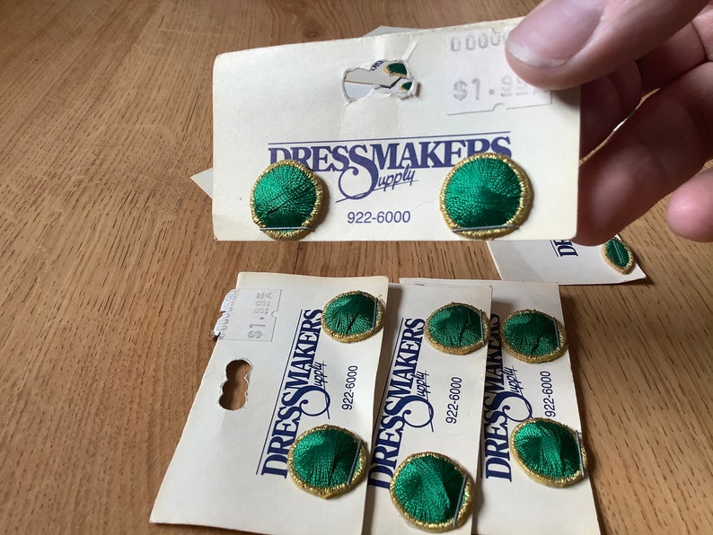 May include: Five green and gold embroidered buttons on white cards with the text "Dressmakers Supply" and the phone number "922-6000".