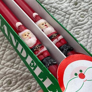 Puede incluir: Dos velas rojas de Santa Claus en una caja verde y blanca. La caja dice "CANDLE" en la parte delantera.