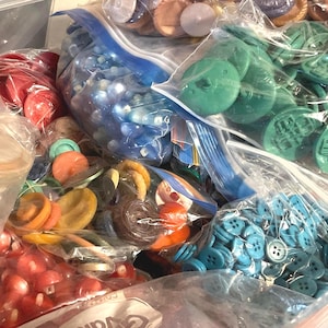 May include: A collection of colorful buttons in various shapes and sizes, stored in clear plastic bags. The buttons are in shades of red, orange, yellow, green, blue, and purple. Some buttons have two or four holes, while others have a shank.