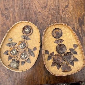 以下が含まれることがあります： 茶色の籐で編まれた楕円形の壁掛け2つ。茶色のココナッツの殻でできた花と白いアクセントが施されています。花は花瓶の形に配置されています。