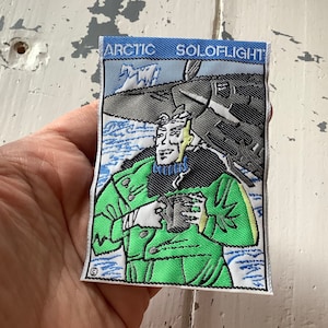 May include: An embroidered patch featuring a cartoon pilot in a green flight suit and a black fur-lined jacket. The patch is green and black with the text "ARCTIC SOLOFLIGHT" in white.
