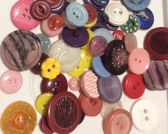 May include: A collection of various buttons in different sizes, shapes, and colors. The buttons include shades of pink, purple, yellow, red, blue, and white. Some buttons have textured surfaces, while others are smooth.