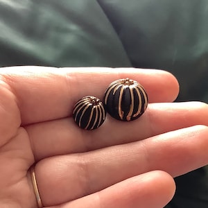 Puede incluir: Dos botones redondos negros y dorados con un diseño decorativo. El botón más grande tiene un diámetro de aproximadamente 2,5 cm y el botón más pequeño tiene un diámetro de aproximadamente 2 cm.