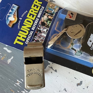 May include: A silver whistle with a black lanyard and a silver Acme Thunderer label. The whistle is packaged with a blue and white card that reads "Official Referee Whistle" and "The Acme Thunderer".