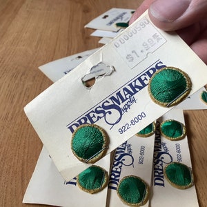 May include: A close-up of a package of green and gold fabric covered buttons. The buttons are labeled "Dressmakers Supply" and have a phone number of 922-6000.