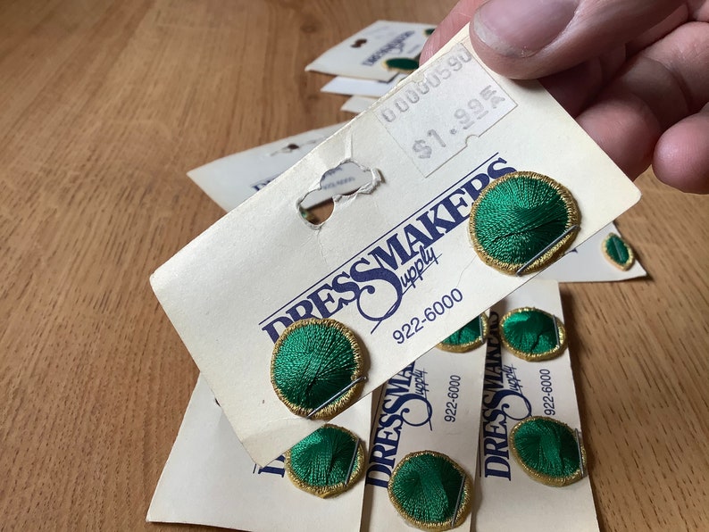 May include: A close-up of a package of green and gold fabric covered buttons. The buttons are labeled "Dressmakers Supply" and have a phone number of 922-6000.
