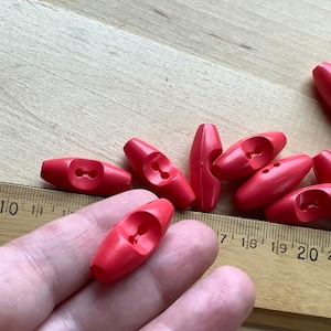 以下が含まれることがあります： 明るい赤色のプラスチック製のトグルボタンが10個あります。ボタンは楕円形で、中央に2つの穴があります。ボタンは木製の表面に積み重ねられています。