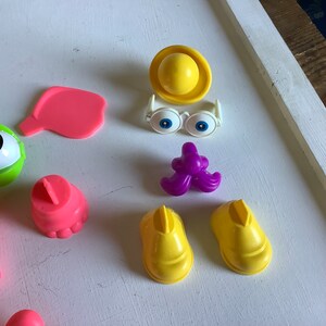 Może przedstawiać: Zestaw plastikowych części do zabawki Potato Head. Zestaw zawiera r&oacute;żowy nos, fioletowy wąs, ż&oacute;łty kapelusz, ż&oacute;łte buty, r&oacute;żowe uszy i białe okulary z niebieskimi oczami.