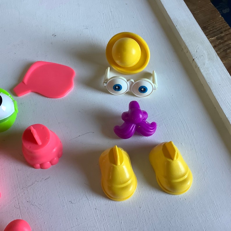 Może przedstawiać: Zestaw plastikowych części do zabawki Potato Head. Zestaw zawiera r&oacute;żowy nos, fioletowy wąs, ż&oacute;łty kapelusz, ż&oacute;łte buty, r&oacute;żowe uszy i białe okulary z niebieskimi oczami.