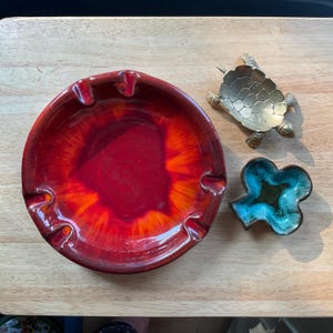 May include: A collection of decorative ashtrays. A large, round, red and orange ashtray sits next to a small brass turtle-shaped ashtray and a turquoise and green clover-shaped dish.
