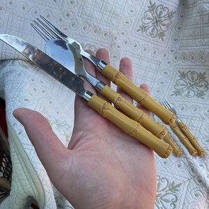 May include: A set of stainless steel silverware with bamboo-style handles. The set includes a knife, spoon, and fork, with additional forks visible. The handles are a light yellow color, and the silverware is held in a hand.