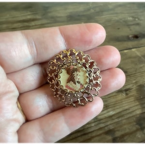May include: A gold-toned brooch with a delicate filigree design. The centre features a small, embroidered portrait of a woman in a pink dress.