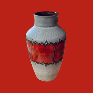 May include: A grey ceramic vase with a red and orange painted band, outlined in black, near the centre. The vase has a wide base, a narrow neck, and a dark brown interior. The background is a solid red.