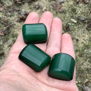 Puede incluir: Tres piedras lisas, de color verde oscuro, pulidas con un patrón estriado. Las piedras tienen forma de rectángulos redondeados.