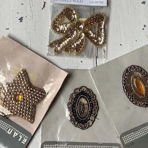 May include: Four gold and black iron-on patches. One is a star shape, one is a bow, one is a round oval, and one is a larger oval with a clear gem in the center.