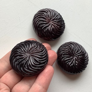 Puede incluir: Tres botones decorativos redondos de color morado oscuro con un diseño de remolino y textura.