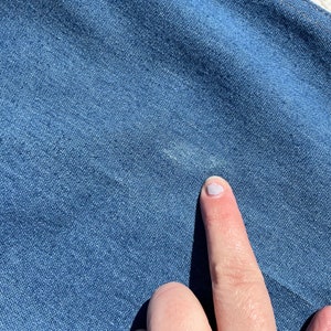 Op de afbeelding: Close-up van een blauwe denim stof met een witte vlek. Een vinger wijst naar de vlek.