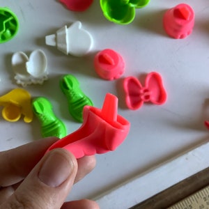 Może przedstawiać: Zestaw kolorowych plastikowych foremek do ciastek w r&oacute;żnych kształtach, w tym but, kokarda, rękawiczka i kapelusz. Formy są r&oacute;żowe, zielone, ż&oacute;łte i białe.