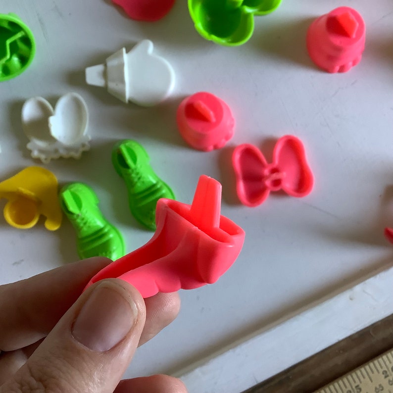 Może przedstawiać: Zestaw kolorowych plastikowych foremek do ciastek w r&oacute;żnych kształtach, w tym but, kokarda, rękawiczka i kapelusz. Formy są r&oacute;żowe, zielone, ż&oacute;łte i białe.