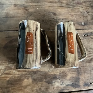 May include: Two ceramic mugs with brown and black abstract designs and gold accents. The mugs have a handle on the side.