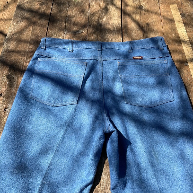Op de afbeelding: Een paar blauwe spijkerbroeken met een achterzak aan elke kant. De broek heeft een bruin leren patch met het woord "Wrangler" erop.