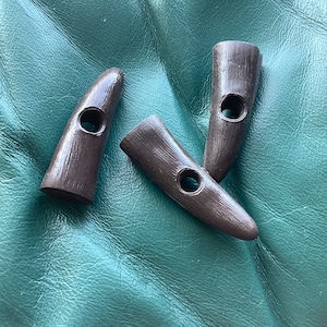 Könnte beinhalten: Drei braune Kunststoff-Hornknöpfe mit einem Loch in der Mitte jedes Knopfes. Die Knöpfe liegen auf einer grünen Oberfläche.