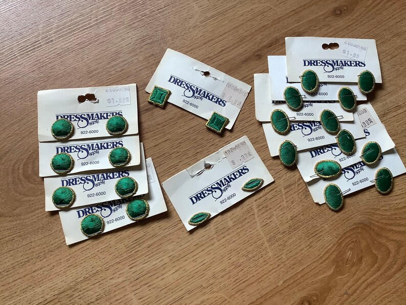 May include: Assortment of green and gold fabric covered buttons with the text "Dressmakers Supply" and a phone number. The buttons are packaged on white cards.