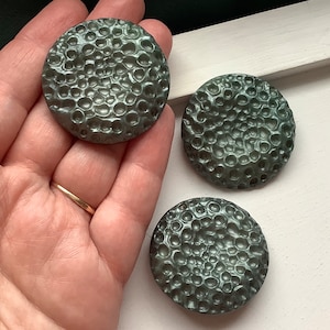 Könnte beinhalten: Drei runde, dunkelgraue, strukturierte Knöpfe mit einer erhabenen, unebenen Oberfläche. Die Knöpfe haben einen leicht metallischen Glanz.