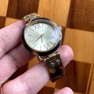 Peut inclure: Une montre dorée avec un bracelet en écaille de tortue. Le cadran de la montre est rond et comporte des chiffres romains pour les heures. Le cadran de la montre indique "Anne Klein".