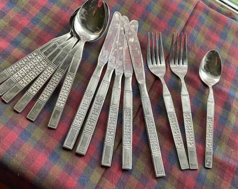 Op de afbeelding: Een verzameling vintage bestek, waaronder lepels, messen en vorken, gerangschikt op een geruite tafelkleed. Het bestek heeft een decoratief patroon op de handvatten. De lepels hebben een afgeronde kom en de messen hebben een gekartelde rand.