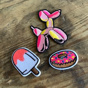May include: Three embroidered iron-on patches: a pink balloon animal dog, a red and white popsicle, and a pink donut with colourful sprinkles.