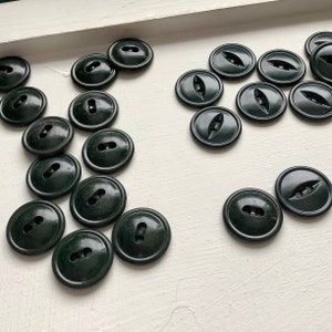 May include: A collection of dark green plastic buttons with two holes. The buttons are arranged in a random pattern on a white surface.