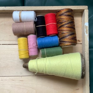 May include: A wooden tray filled with spools of colourful thread, including yellow, pink, green, blue, red, brown, white, and black. There is also a large spool of light green thread.