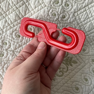 Może przedstawiać: Czerwony plastikowy hak z dwoma pętlami. Hak jest przeznaczony do zawieszania przedmiotów na powierzchni.