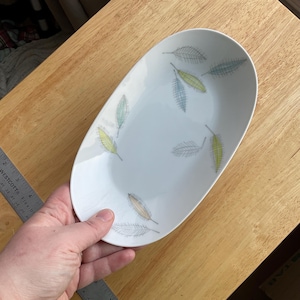 Op de afbeelding: Witte ovale serveerschaal met een bladerpatroon in groen, geel en grijs. De schaal wordt door een hand vastgehouden en ligt op een houten oppervlak. De schaal is ongeveer 30 cm lang.