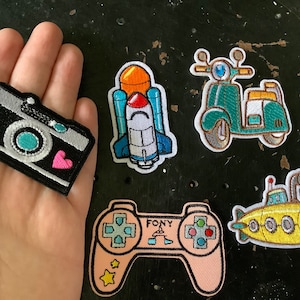 May include: A selection of embroidered patches including a camera, rocket, scooter, game controller, and submarine. The patches are colourful, with a black camera, a blue and white rocket, a teal scooter, a pink controller, and a yellow submarine.