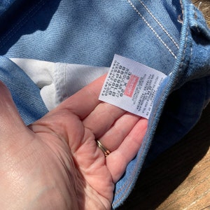 Op de afbeelding: Een blauw denim jean label met witte stiksels. Het label is bevestigd aan de binnenkant van de jean en heeft het Wrangler logo, maat 36 x 29, Regular Fit en een stylenummer. Het label is gemaakt in Nicaragua.