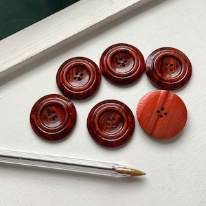May include: Six brown wooden buttons with four holes each. The buttons have a reddish-brown colour and a striped pattern.