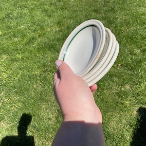 May include: A stack of four white ceramic plates with a green trim. The plates are being held in a person's hand.
