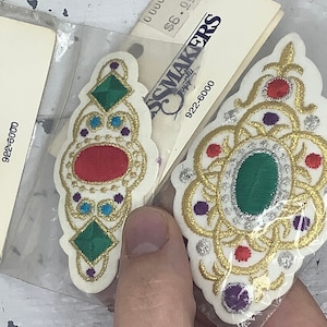 May include: Two white fabric patches with gold embroidery. One patch features a red oval gem surrounded by smaller gems in purple, blue, and green. The other patch features a green oval gem surrounded by smaller gems in red and purple. Both patches have the text "Dressmakers Supply 922-6000".
