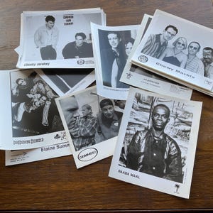 Puede incluir: Una colección de fotografías promocionales en blanco y negro para varios artistas musicales. Las fotografías están apiladas sobre una superficie de madera. Los artistas incluyen Chewy Marble, Baaba Maal, Church Gun Terror, Hot House Flowers, Elaine Summers e Iren Hoffman.
