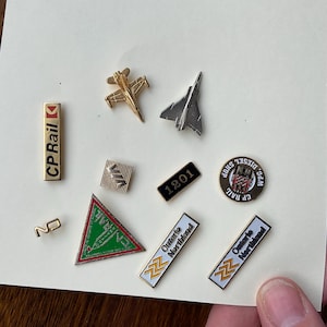 Op de afbeelding: Een verzameling van negen emaillepins met verschillende ontwerpen, waaronder een straalvliegtuig, een trein en een logo voor Ontario Northland. De pins bevinden zich op een wit oppervlak.