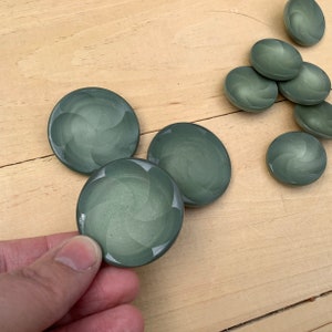 Könnte beinhalten: Ein Satz von acht grünen runden Knöpfen mit einem Wirbelmuster. Die Knöpfe sind aus einem glatten, glänzenden Material gefertigt.