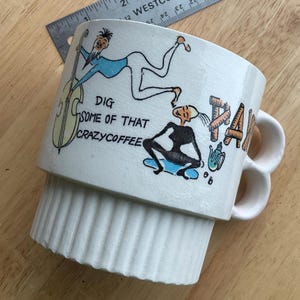 Puede incluir: Taza de café de cerámica blanca con un diseño apilado único y dos asas. La taza presenta una ilustración caprichosa con las palabras "DIG SOME OF THAT CRAZYCOFFEE" y un diseño temático de jazz con figuras de dibujos animados e instrumentos musicales.
