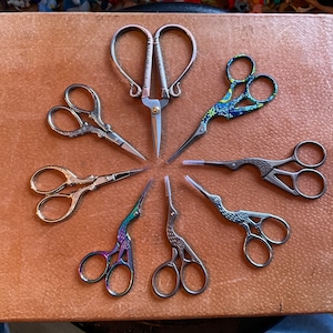 May include: A collection of eight decorative scissors with various designs and finishes. The scissors are arranged in a circular pattern on a brown surface.
