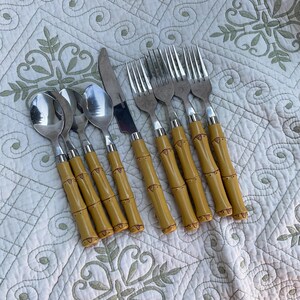May include: A set of stainless steel flatware with bamboo-style handles. The set includes spoons, forks, and knives. The handles are a light yellow color with dark brown accents, and the utensils are arranged on a patterned white surface.