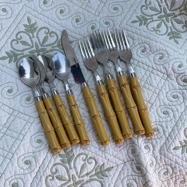 May include: A set of stainless steel flatware with bamboo-style handles. The set includes spoons, forks, and knives. The handles are a light yellow color with dark brown accents, and the utensils are arranged on a patterned white surface.